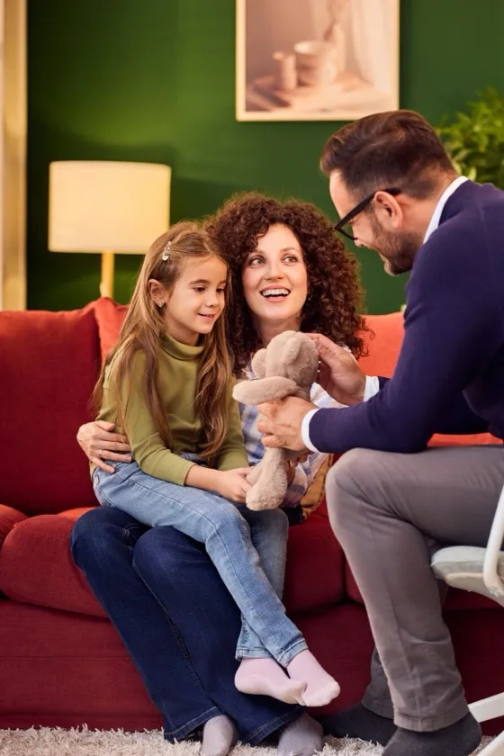 A child holds a teddy bear close while a parent sits beside them at home.