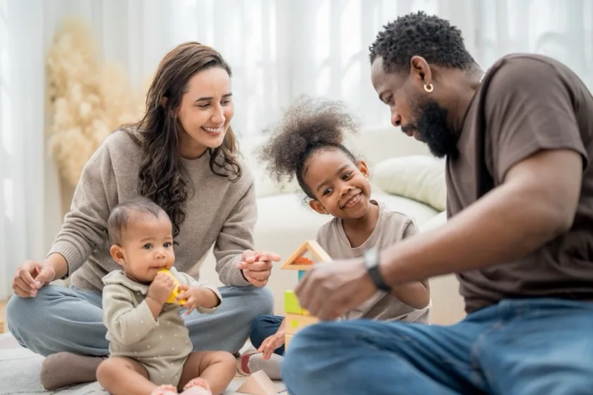Two parents sit on the floor stacking blocks with their toddler.